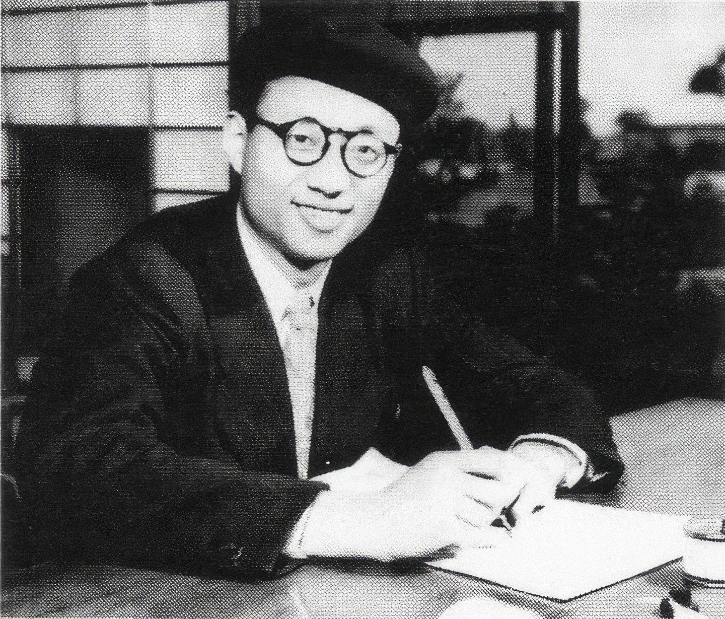 Black and white photo of a smiling man named Osamu Tezuka sitting at a table, wearing a beret and glasses. He is writing on a piece of paper with his right hand, and his left hand appears to be holding another paper or book down on the table.